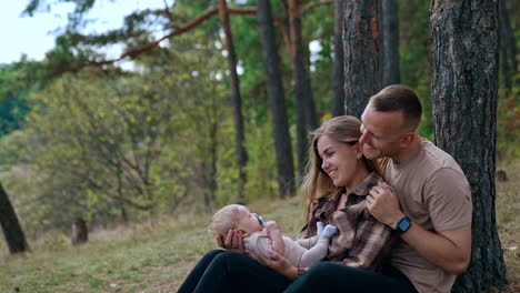 man embraces his wife holding a baby boy. caucasian couple sits near the tree smiling to their child with pacifier.