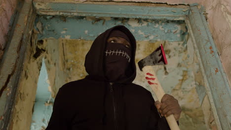 bottom view of man wearing black balaclava and holding an ax in a ruined building