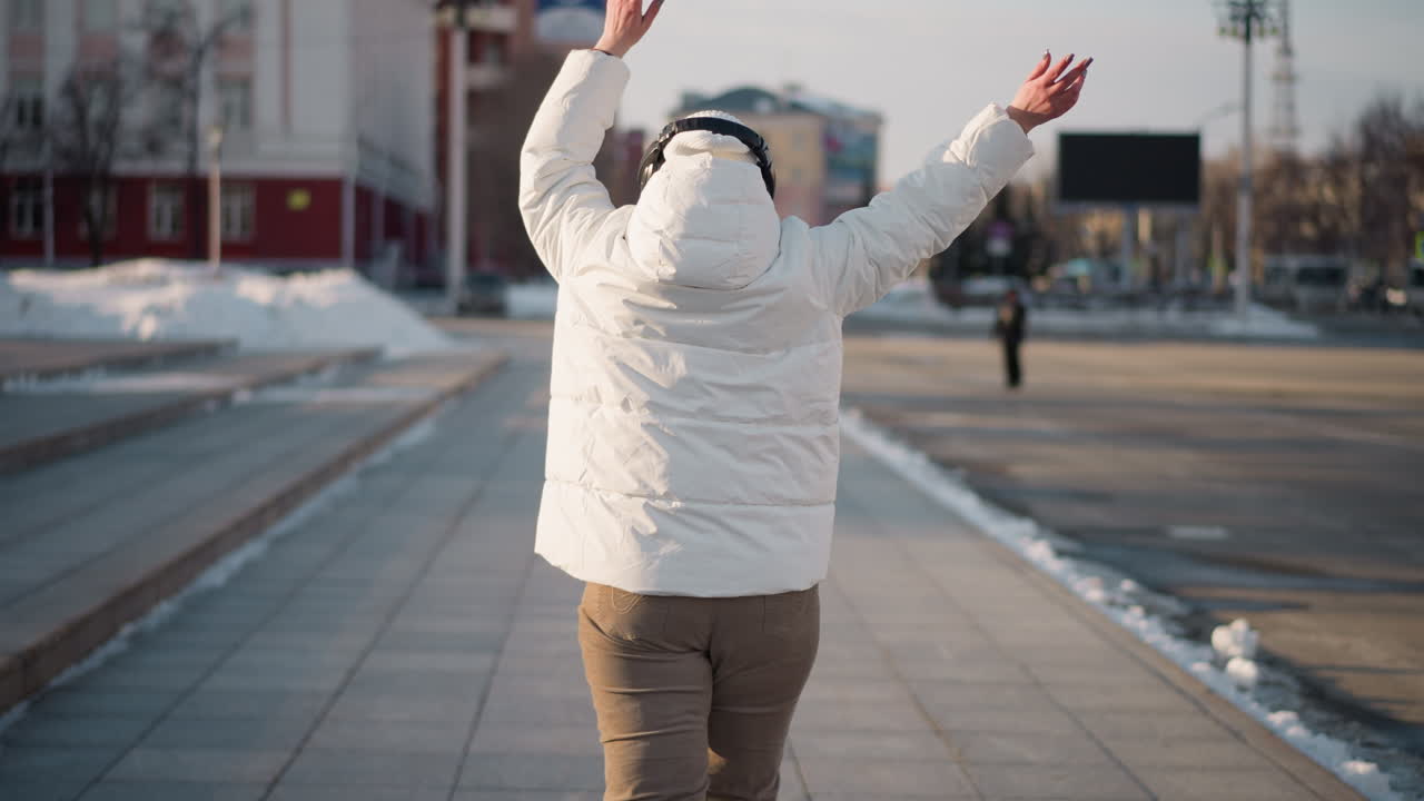 Back view of snow dancer grooving along tiled walkway in urban winter street, arms outstretched in motion while wearing puffy white jacket, beanie, and headphones, surrounded by snow, and billboards