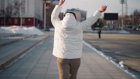 back view of snow dancer grooving along tiled walkway in urban winter street, arms outstretched in motion while wearing puffy white jacket, beanie, and headphones, surrounded by snow, and billboards