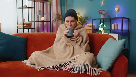 Ill-sick-unhealthy-woman-freezes-from-the-cold-feels-bad-covered-by-warm-blanket-drinks-tea-at-home