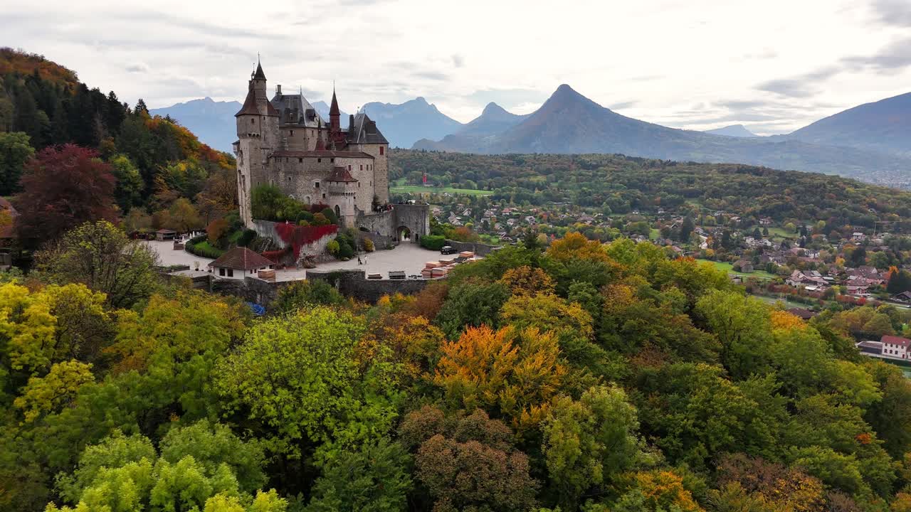 Wide drone establishing shot of Château de Menthon‑Saint‑Bernard and bourg above the forests and villages on the eastern side of Lake Annecy, with the Bauges and Bornes massifs in the background