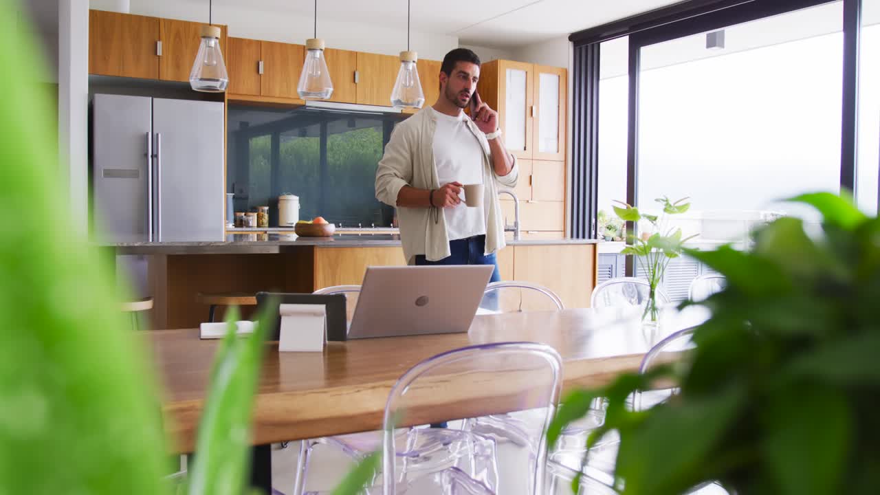 Uomo adulto che risponde a una chiamata in arrivo sullo smartphone mentre tiene una tazza, guardando il laptop in cucina per lavoro.