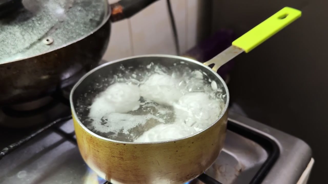 Nærbilde ovenfra av hvite egg som koker i en liten kjele med vann på en gasskomfyr. Detaljerte bobler og damp under matlaging på kjøkkenet. Ideelt for innhold om matlaging og sunn livsstil.
