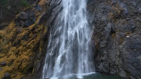 Close-up-and-tilt-up-aerial-shot-of-a-dramatic-waterfall-cascading-down-a-rugged-cliff-surrounded-by-the-natural-rugged-terrain-and-lush-greenery