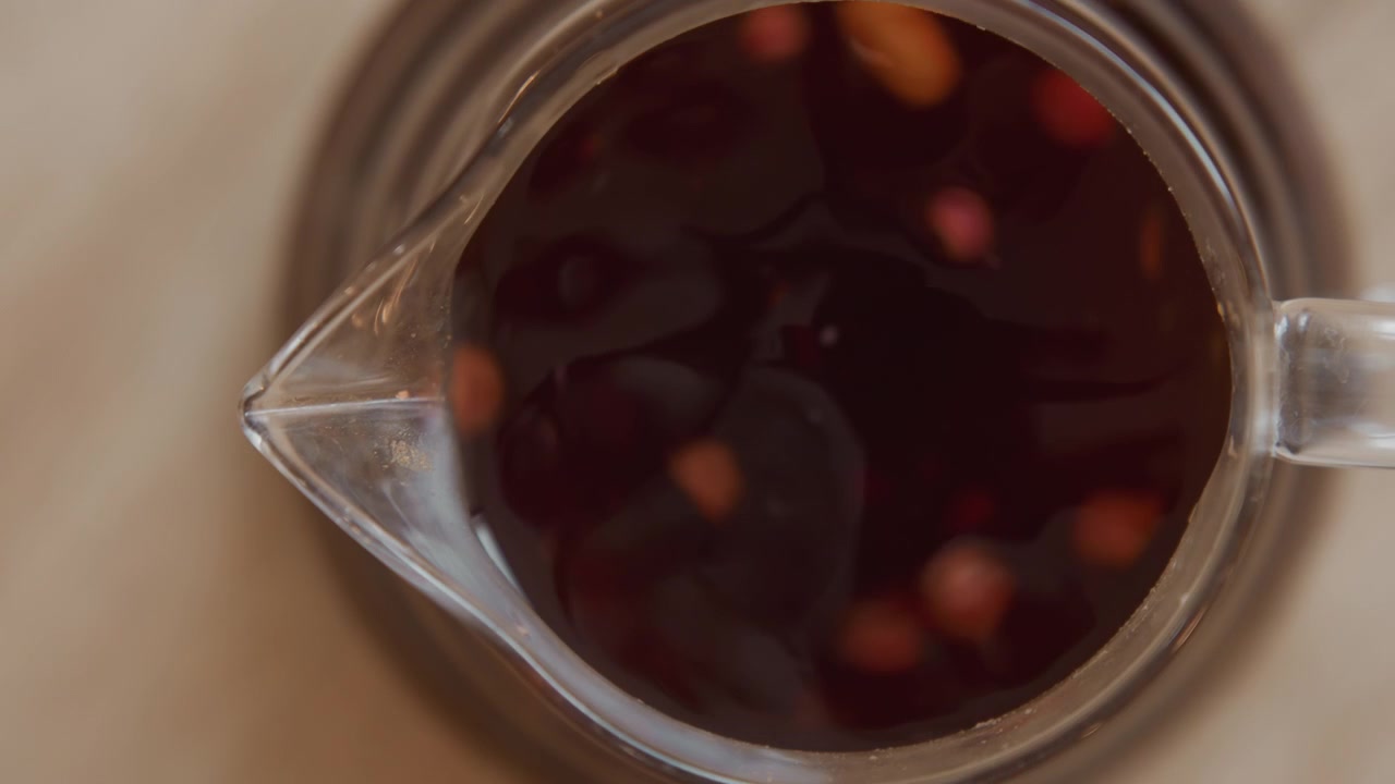 Primo piano di liquido scuro con particelle rosse e arancioni fluttuanti in un contenitore di vetro trasparente. Il livello del liquido sale costantemente mentre viene versato altro contenuto, mostrando dettagli della texture superficiale liscia.