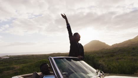 Diverse-couple-driving-on-sunny-day-in-convertible-car-woman-standing-raising-her-arms-in-the-air