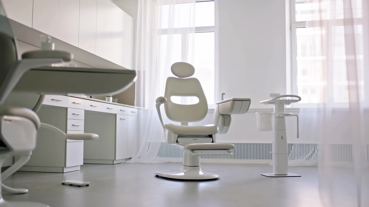 Studio dentistico vuoto e pulito centrato su una poltrona da trattamento ergonomica con poggiatesta regolabile, braccioli, unità laterale, sgabello per assistente e comandi a pedale, illuminato da luce diurna soffusa in un interno luminoso e sterile.