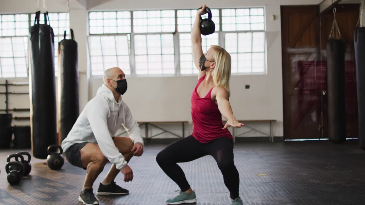 mujer caucásica en forma con máscara facial trabajando con campanas de hervidor con entrenador masculino en el gimnasio