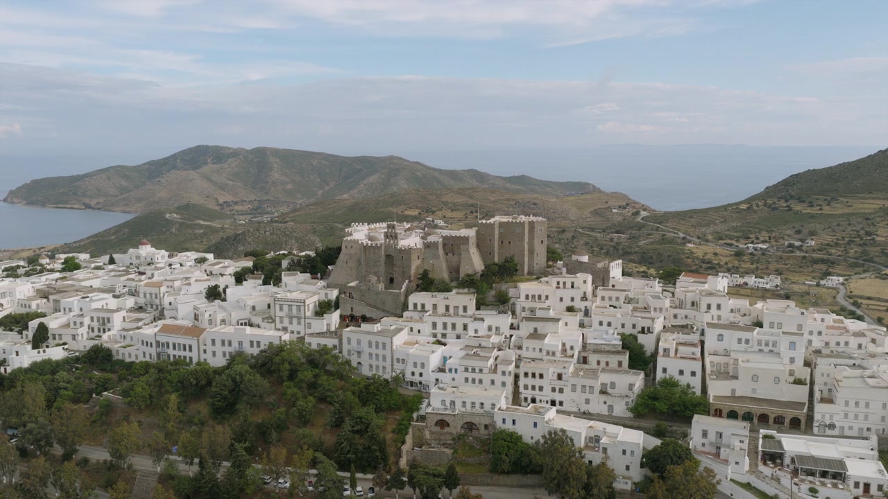 Vista aérea del pueblo de Chora en Patmos y el Monasterio fortificado de San Juan, enmarcado por colinas ondulantes y el mar distante.