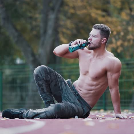 handsome athlete drinks water outdoors. strong shirtless man with muscular body sitting on the stadium track and resting after his workout.