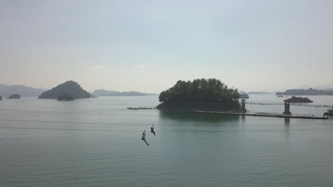 aerial pan follows tourists sliding down zip line over lake in china