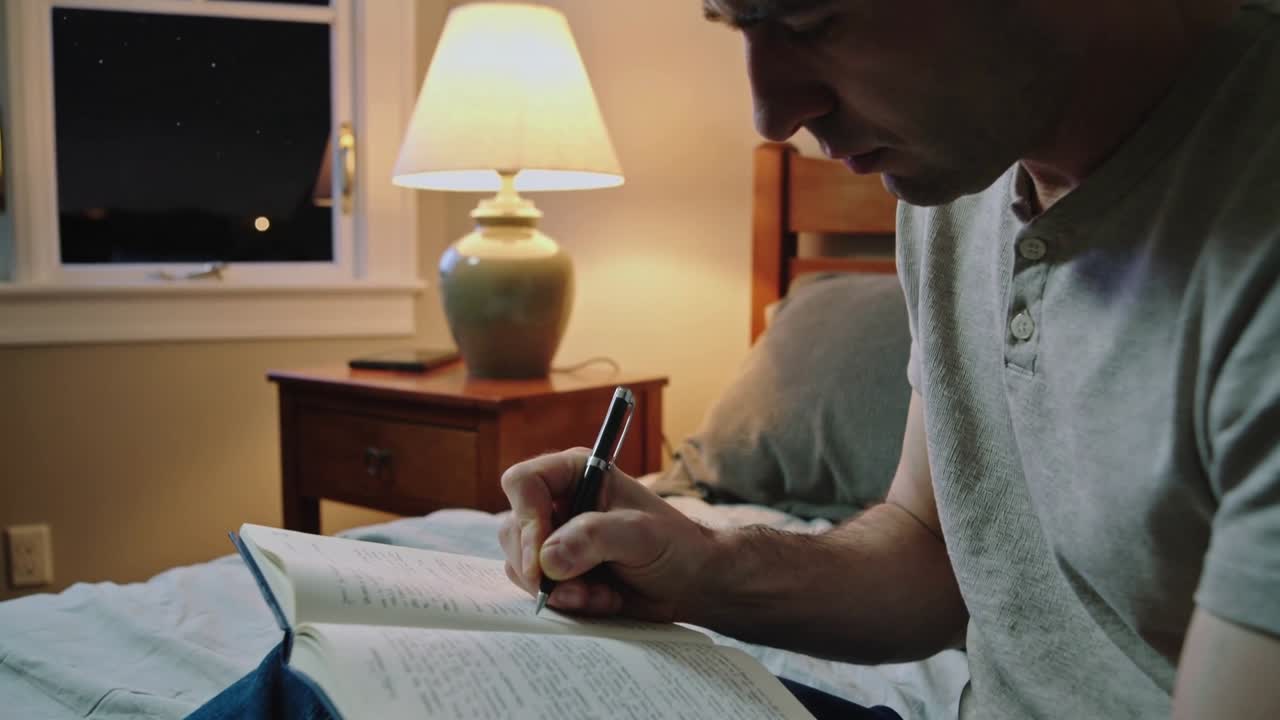 Uomo che scrive, indossando una maglietta henley grigia, con una penna sospesa sopra un quaderno sul letto, che scrive un diario alla luce di una lampada.