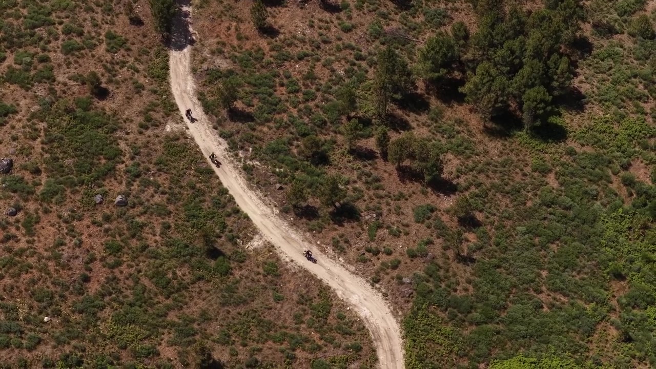 Aerial footage shows several motorcyclists navigating a winding dirt road through open, sunlit terrain with scattered bushes and trees. Clear skies highlight the adventurous, free-spirited mood.