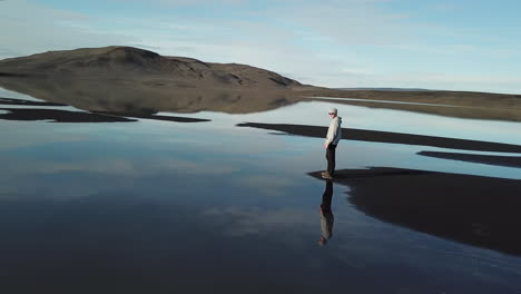 aerial view of lonely man in surreal landspace of iceland highlands, standing by lake with mirror sky reflection