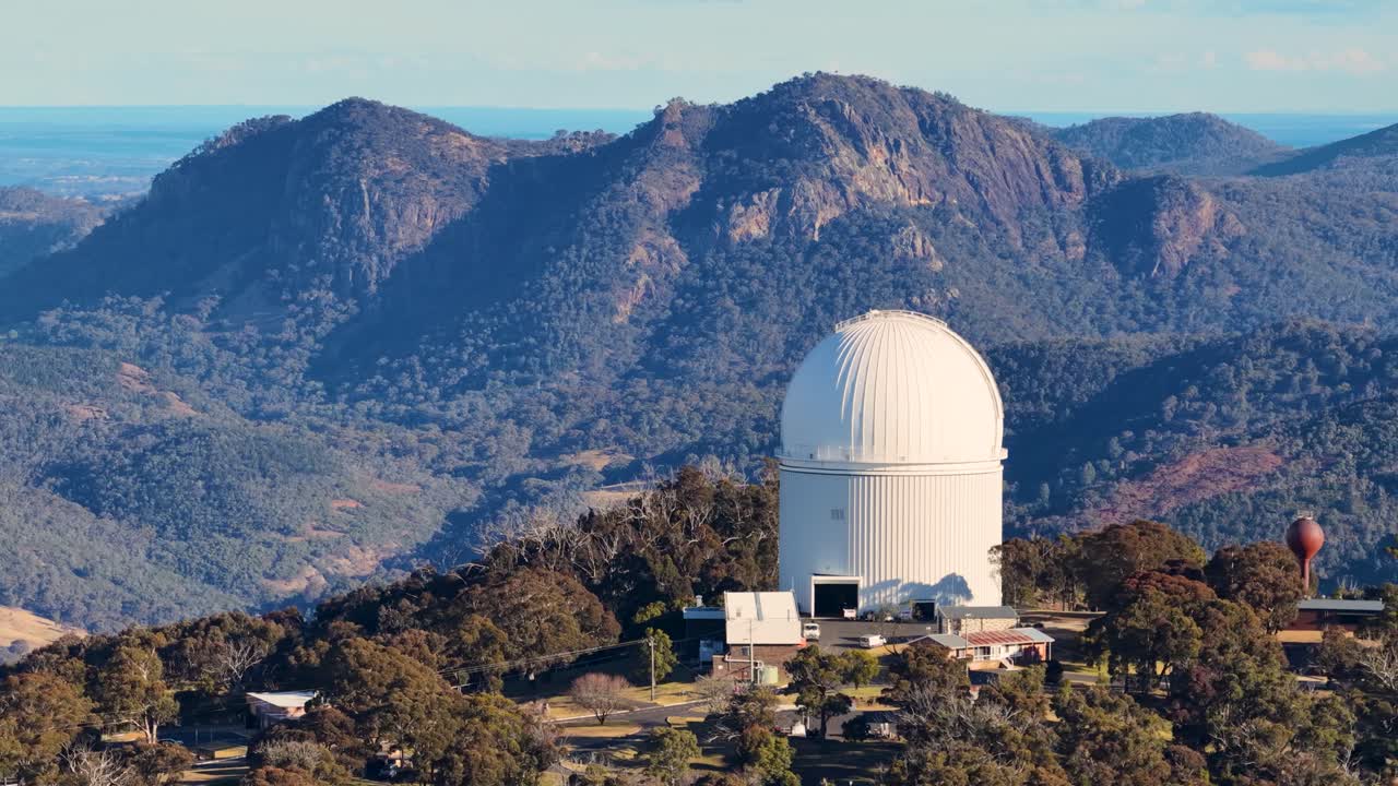 Aerial drone footage orbits a large white observatory dome amid forested hills and rugged mountains during golden hour, with smooth camera movement and natural lighting