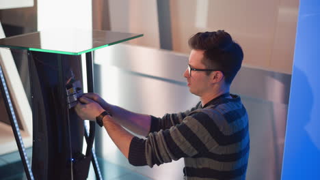 man adjusts display screen setup in video production studio, working on connecting cables and devices, focused on configuration, wearing striped sweater, with blue lights and modern equipment background