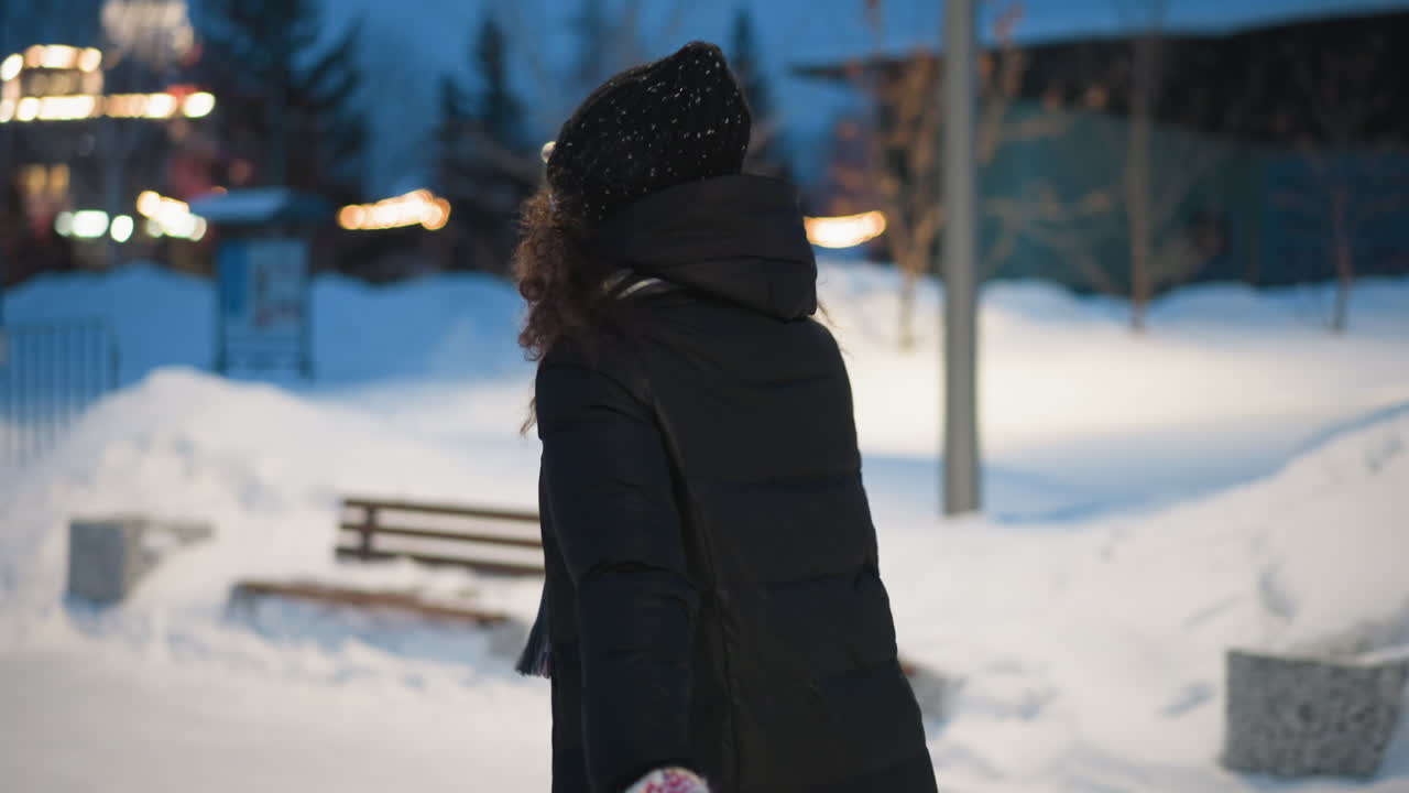 Skater in black winter coat, knitted hat, and colorful mittens glides outdoors on snowy path at dusk with festive blurred lights in background, enjoying cold weather atmosphere with joyful motion