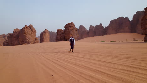 hombre caminando en el desierto del sáhara con una gran formación de piedra arenisca en el fondo en el parque nacional de tassili n'ajjer, argelia