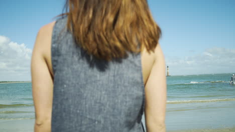 female model walks away towards ocean at beach, lighthouse in distance