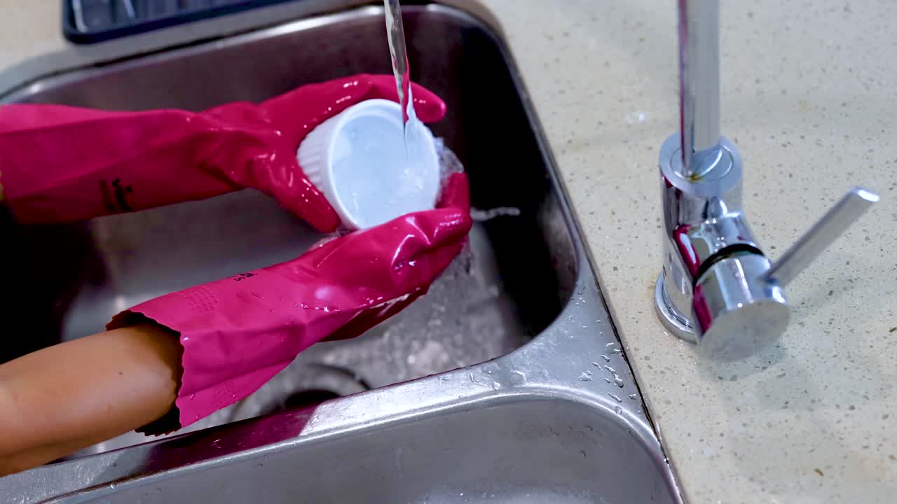 Hands in pink gloves wash a bowl under running water in a kitchen sink. Bright lighting enhances the clean, domestic setting