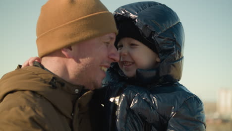 a joyful father carries his playful son close to his ear, with both of them smiling warmly, the child wears a black jacket, while the father wears a brown jacket and a beanie