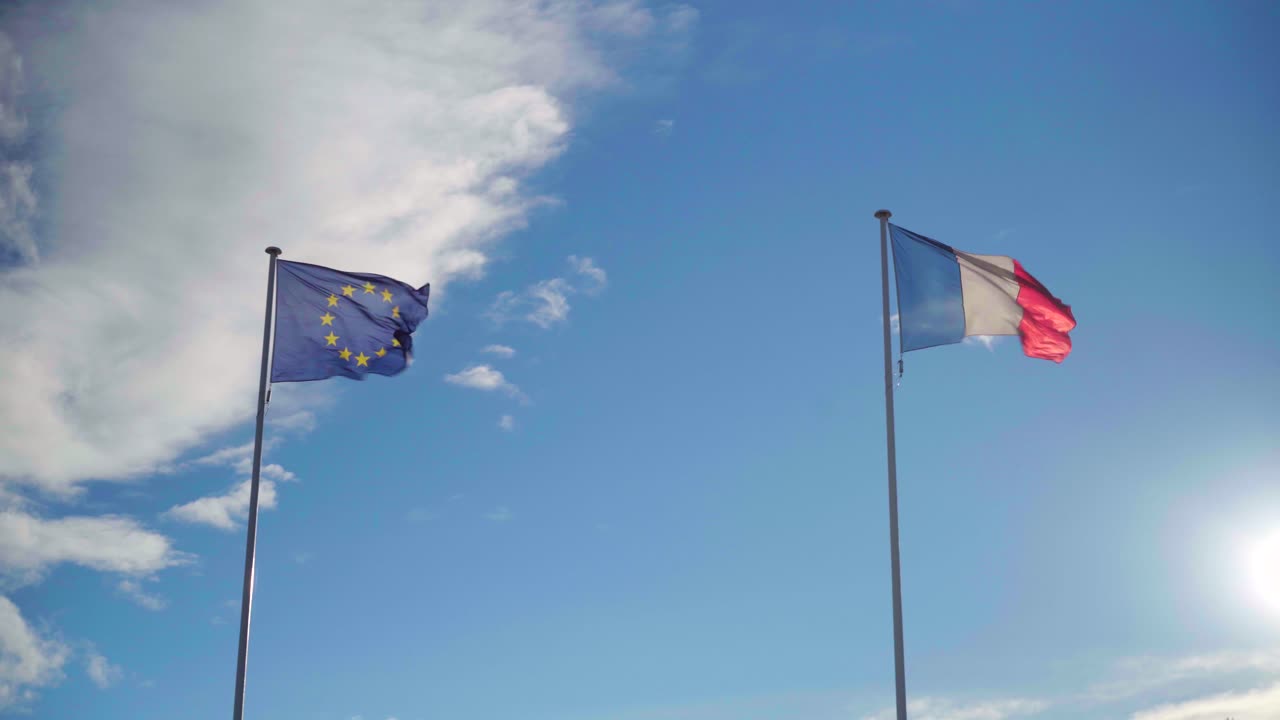 Bandiere europea e francese che sventolano con forte vento su un cielo blu e nuvoloso, simbolo dell'alleanza con l'Unione Europea.