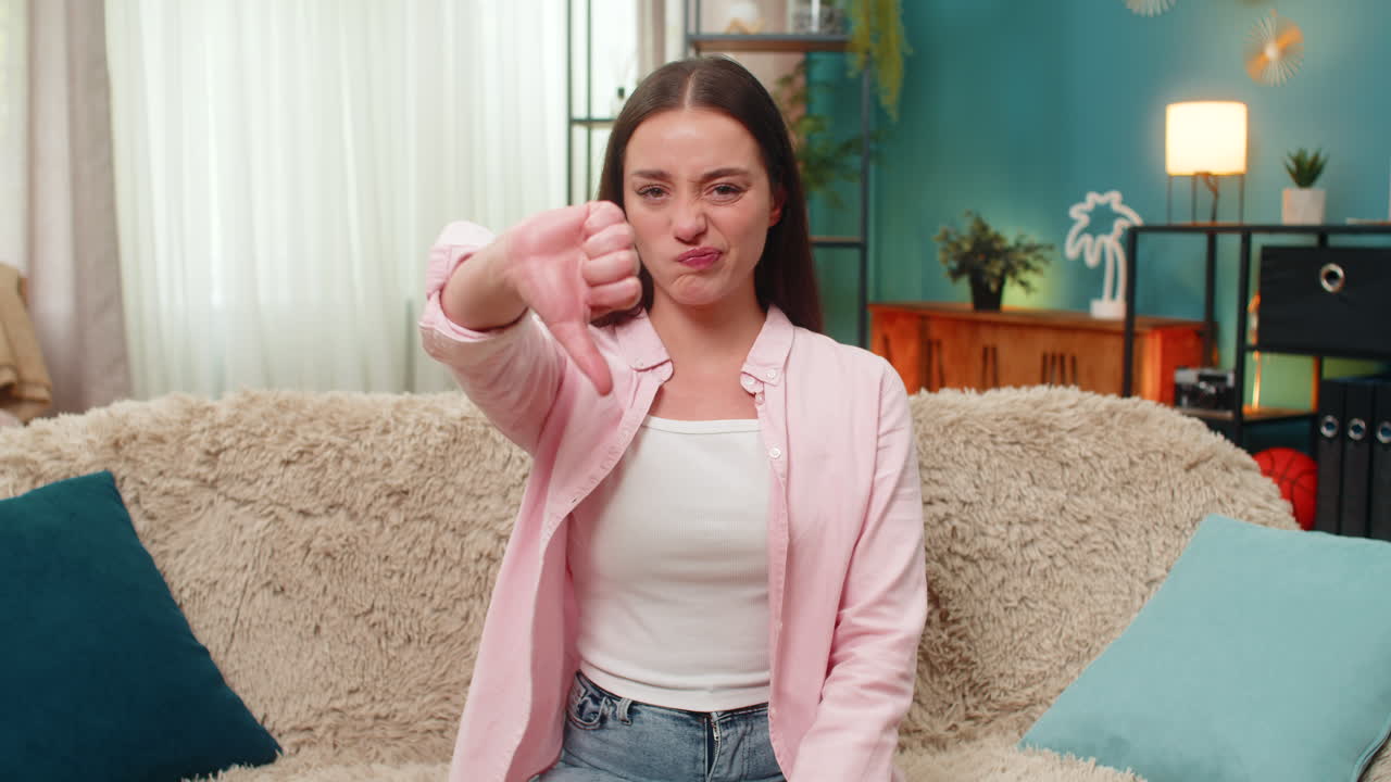 Young woman giving thumbs down gesture frowning showing displeasure and disapproval clearly on sofa