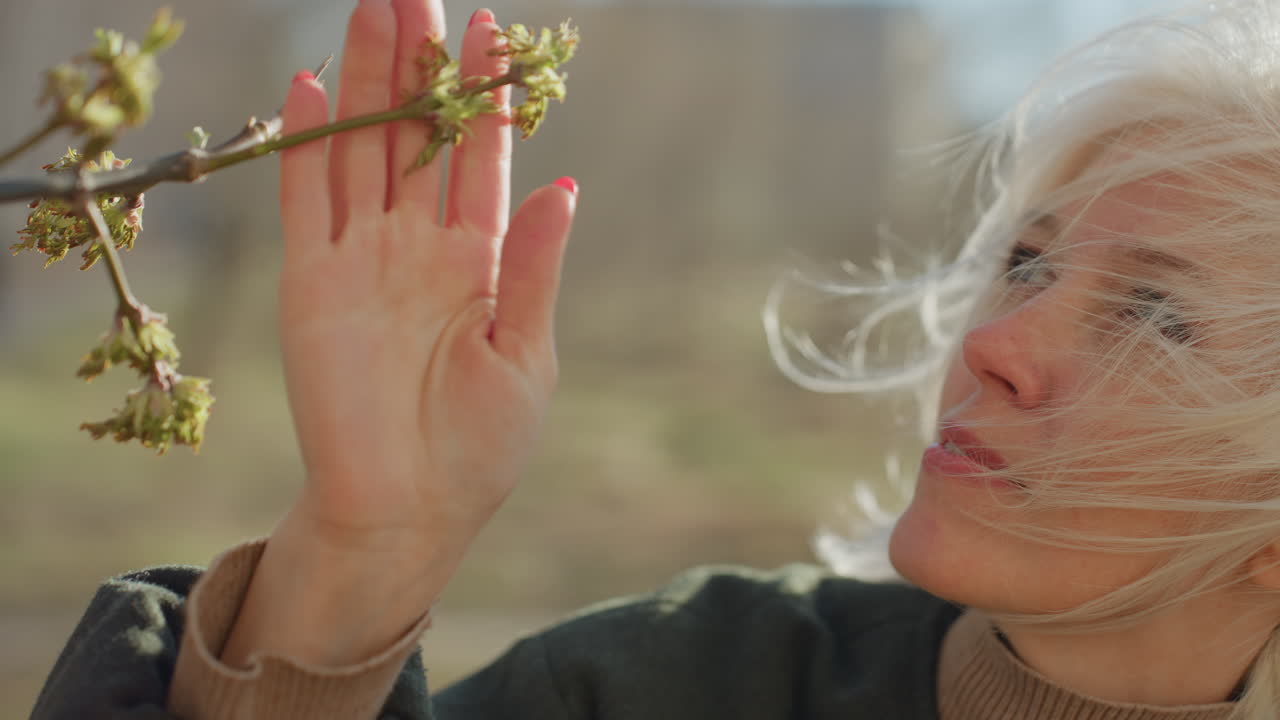 Blonde woman with short hair wearing casual jacket and turtleneck examines budding tree branches with thoughtful expression on sunny spring day, holding twigs gently