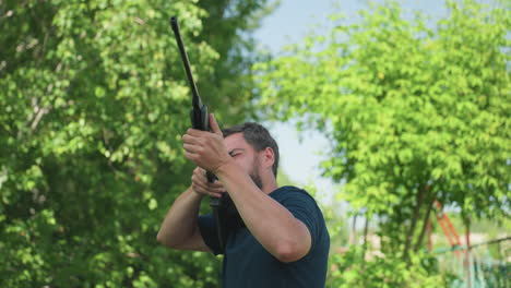 young man reloads pneumatic rifle and aims at unseen target while trees sway in wind, summer rural garden backdrop, cautious wildlife protection and focused preparation for pest control