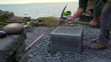 handheld wide shot of a craftsman shaping a square block go cancagua stone with a stone pick, on the shoreline of the city ancud, chiloe island