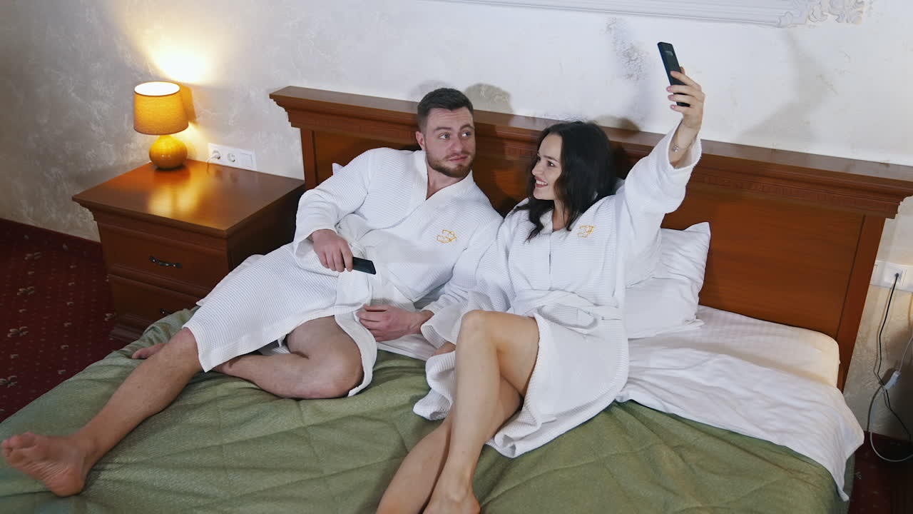 Happy couple in hotel. Young married couple in white bathrobes laying on bed together and doing selfie. Honeymoon.
