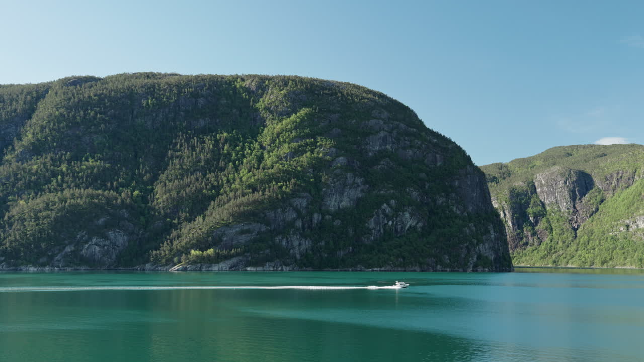 A boat sails across the turquoise-blue waters of Lysefjord in Norway in summer. In the background is a large, steep mountain covered with trees stretching up into the clear blue sky