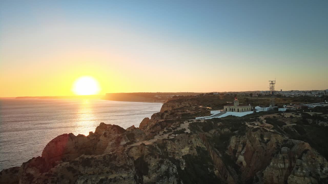 Camera pulls back over dramatic coastal cliffs and Ponta da Piedade lighthouse, revealing sunlit rocks, shimmering waters, and a vibrant sunset along the Algarve shore