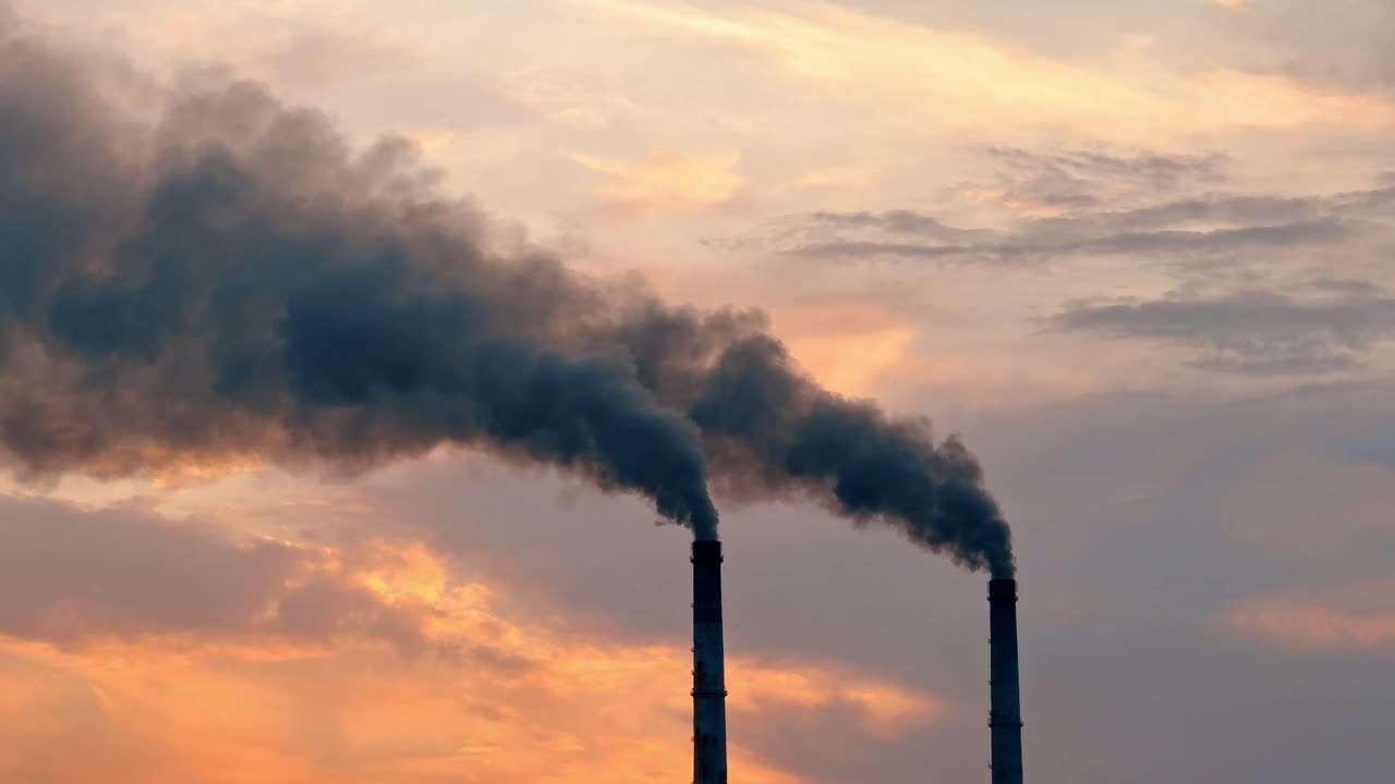 Il fumo si alza da due ciminiere industriali stagliate contro un cielo al tramonto. Fumo scuro si sprigiona da due alte ciminiere mentre il sole tramonta sullo sfondo