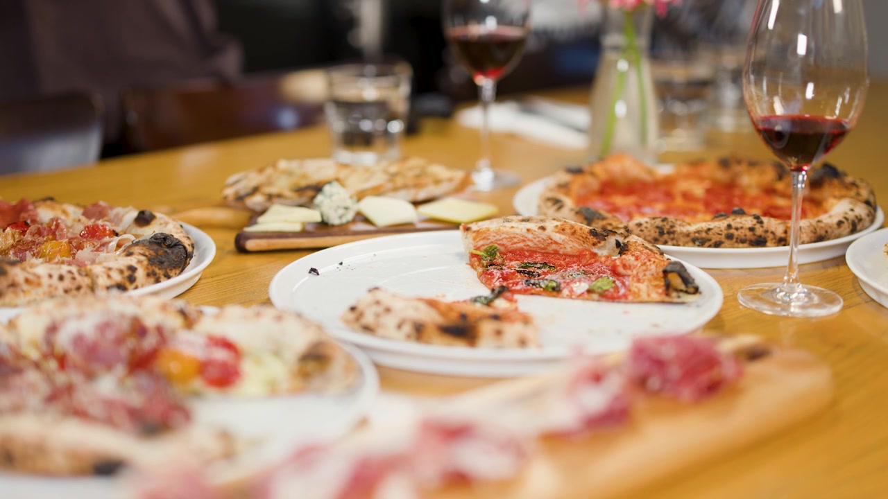 Close-up of hands taking pizza slices from a table with wine and charcuterie appetizers.