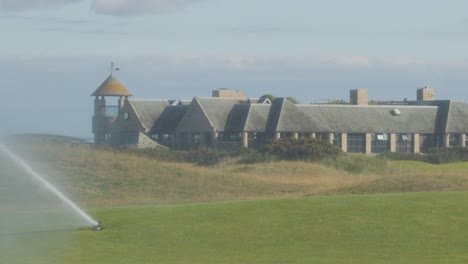 a large sprinkler irrigates a golf course green in front of a clubhouse under daylight, with steady camera and soft natural lighting