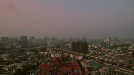 Aerial-rise-up-view-of-Bangkok-city-at-sunset-with-red-rooftops-of-Buddist-temple