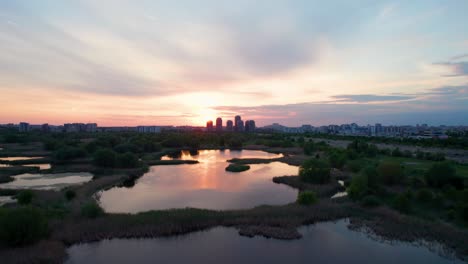 Side-Movement-Drone-View-Over-Vacaresti-Delta-At-Sunset-With-Birds-Flying,-Apartment-Buildings-In-The-Background,-Sunset-Colors,-Orange,-Red,-Yellow,-Bucharest,-Romania