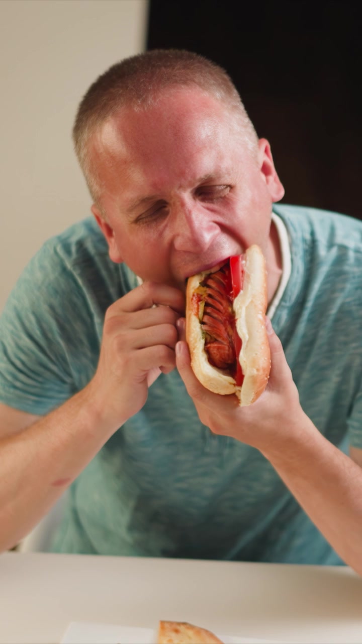 Close up of man eating hot dog with toppings at table. Hot dog bun filled with sausage ketchup and vegetables is held near mouth. White tabletop visible in foreground