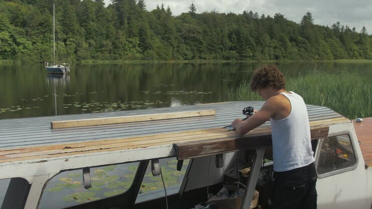 Young man removes masking tape after sealing wooden boat roof starboard side