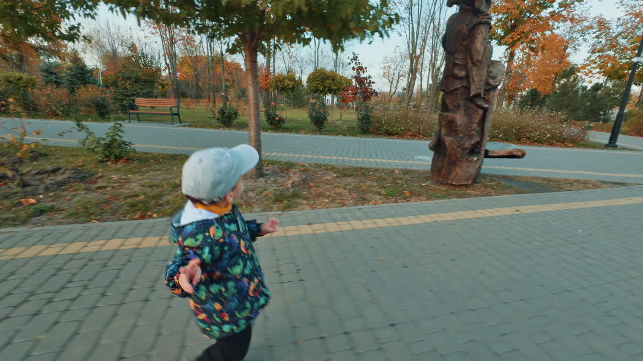 Happy excited baby boy runs cheerfully by the road. Kid stops when notices wooden sculpture in the park.