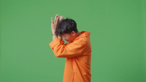 녹색 스크린 배경에 서서 손을 고 있는 아시아 남성 수감자의 측면 뷰, 카메라에 의해 포착되지 않기를 원하는 표정을 보여주는 손바닥 표시