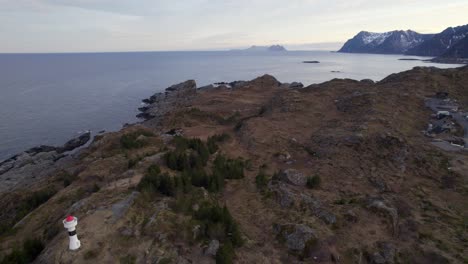 Aerial-backwards-shot-of-a-old-lighthouse-in-Northern-Norway-Lofoten-with-snowy-mountains-in-the-background-and-view-to-Varoy