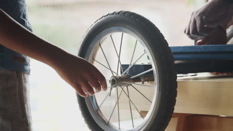 Mid-section-of-pre-teen-boy-checking-the-wheels-of-his-racing-kart-on-a-workbench-while-his-father-attaches-the-frame,-selective-focus