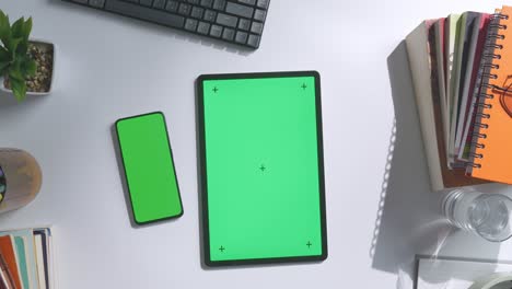 top down view of a laptop computer with mock up green screen chromakey display on a wooden office desk next to notebook with pens, glasses, and a glass of water. slow zoom out, close up