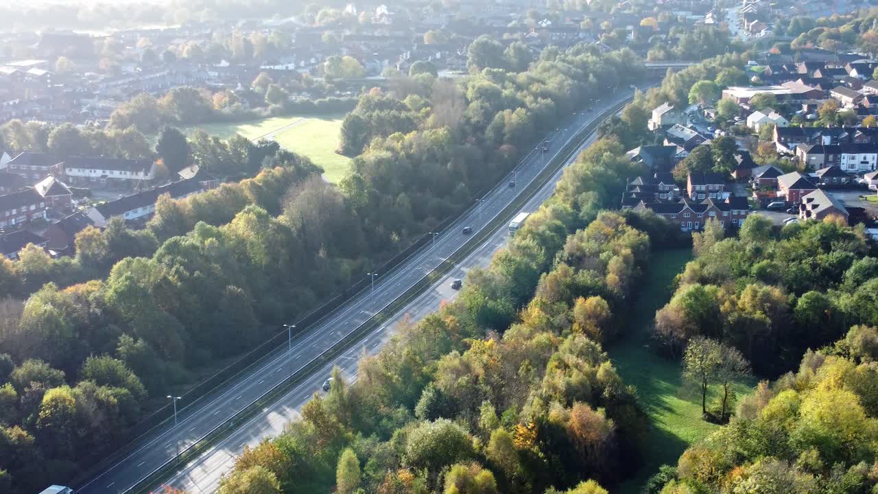 Aerial view tracking across countryside dual carriageway traffic country road between woodland treetop at sunrise towards town