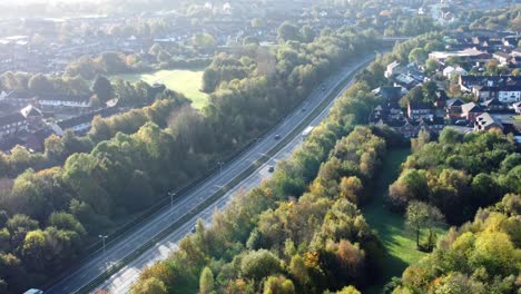 aerial view tracking across countryside dual carriageway traffic country road between woodland treetop at sunrise towards town