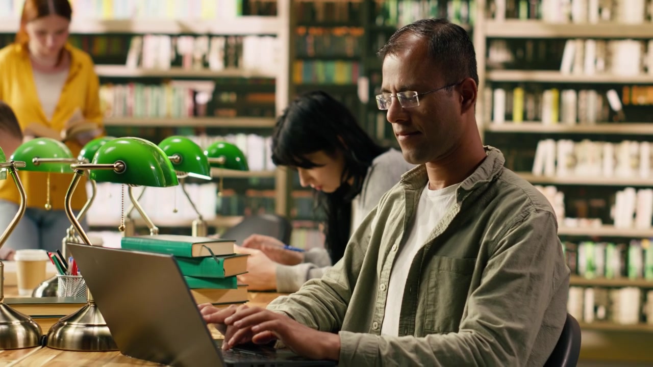 Adult student reading book at a wooden library table with green lamps