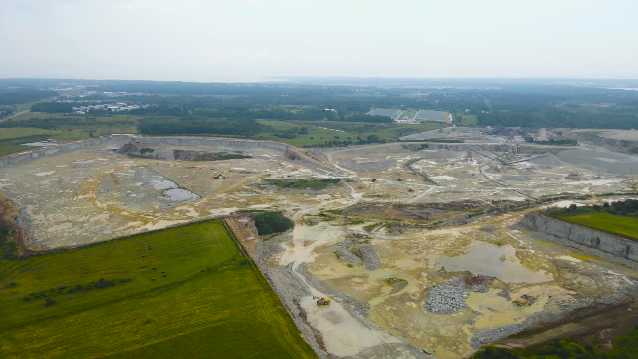 Aerial drone footage flying over a large limestone and gravel quarry mine during a cloudy day in Harku Estonia with green farmfields around. The mine is wet and damp and heavy machinery is visible